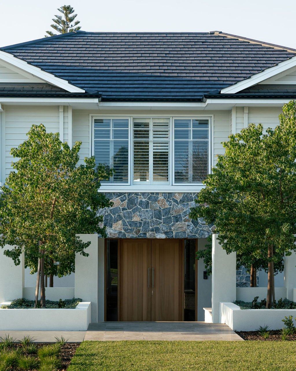 Timber double entry doors under stone cladding, framed by rendered planters and paired trees