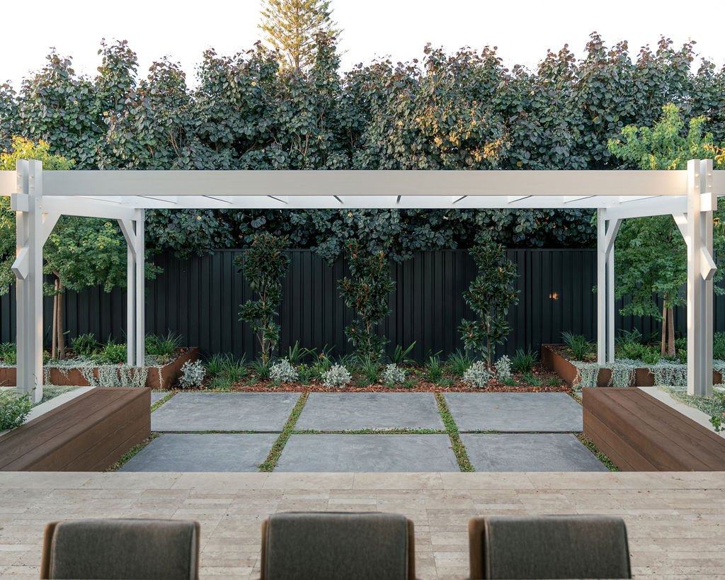 Pergola courtyard with bench seating, pavers and a planted bed against a dark fence
