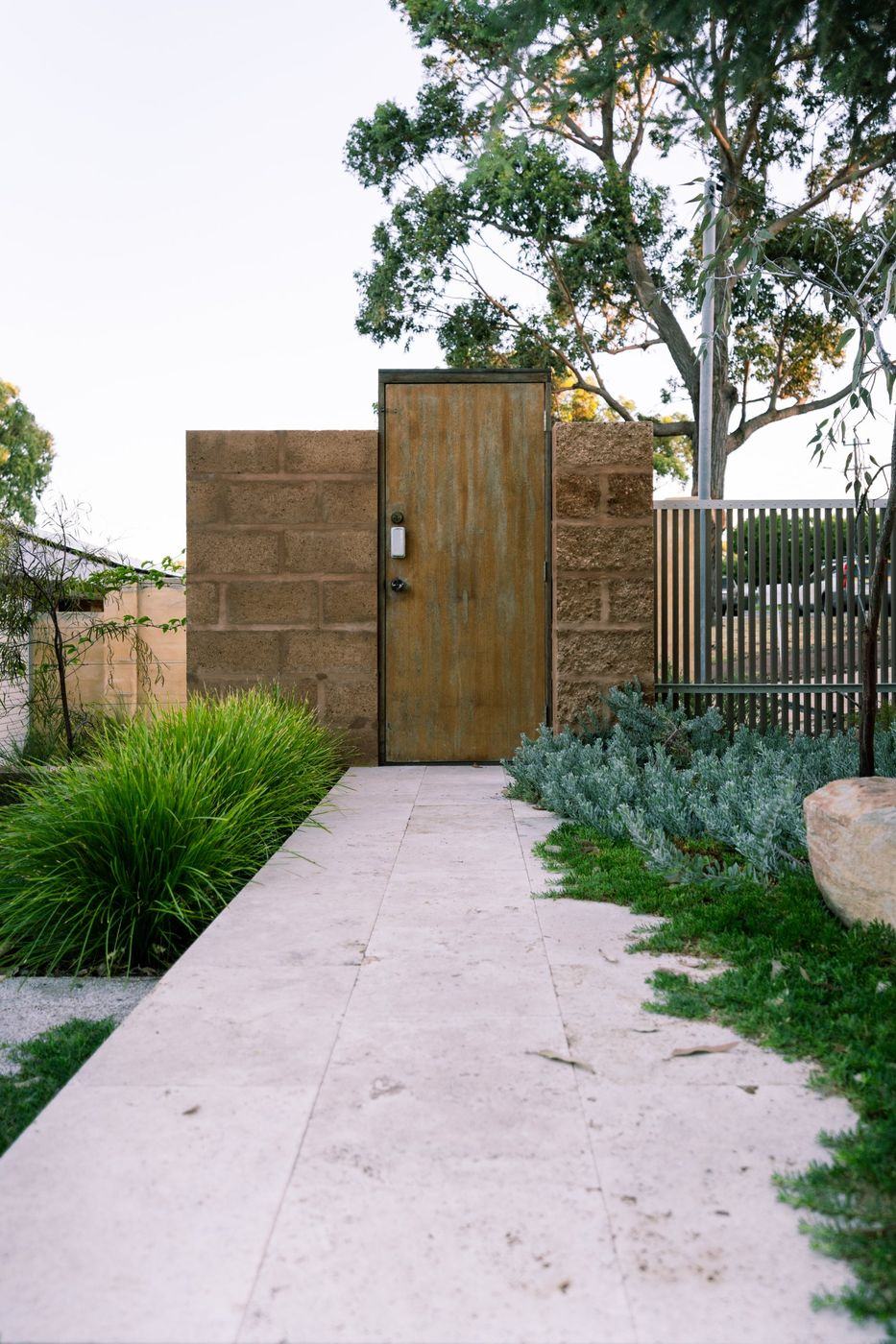 Stone-paved path leading to a weathered timber gate set in a block wall, with grasses and silver groundcover