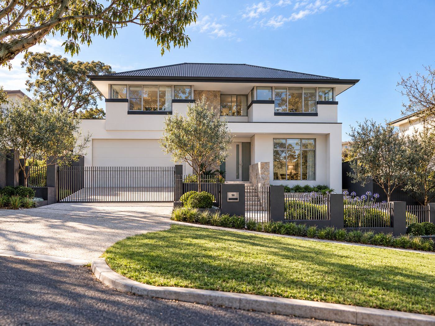 Two-storey white residence with a dark hipped roof, stone feature wall, black palisade fencing, olive trees and agapanthus